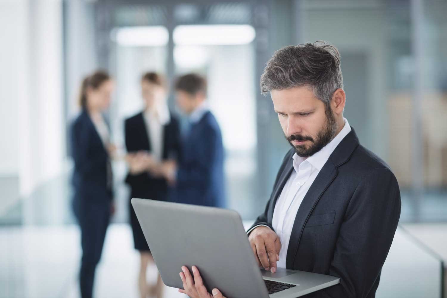 Bearded businessman in a suit looking down at a laptop, with three blurry colleagues talking in the background.
